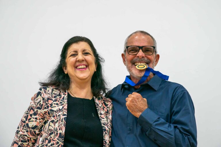 Fotografia destaca, à direita, o professor Waldyr Mendes Ramos, homenageado com a Medalha Minerva do Mérito Acadêmico. À esquerda, a vice-reitora da UFRJ, Cássia Turci. Os dois sorriem e o professor Waldyr faz o popular gesto de morder a medalha.