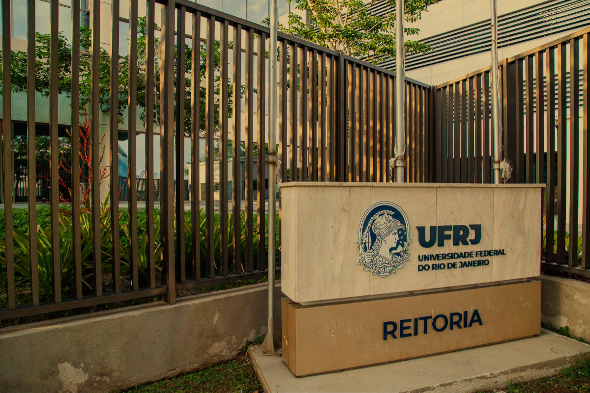 Fotografia mostra, através de grades, parte da fachada do prédio da Reitoria da UFRJ. À frente, há um painel horizontal com a marca completa da Universidade e um letreiro, no qual se lê "Reitoria".