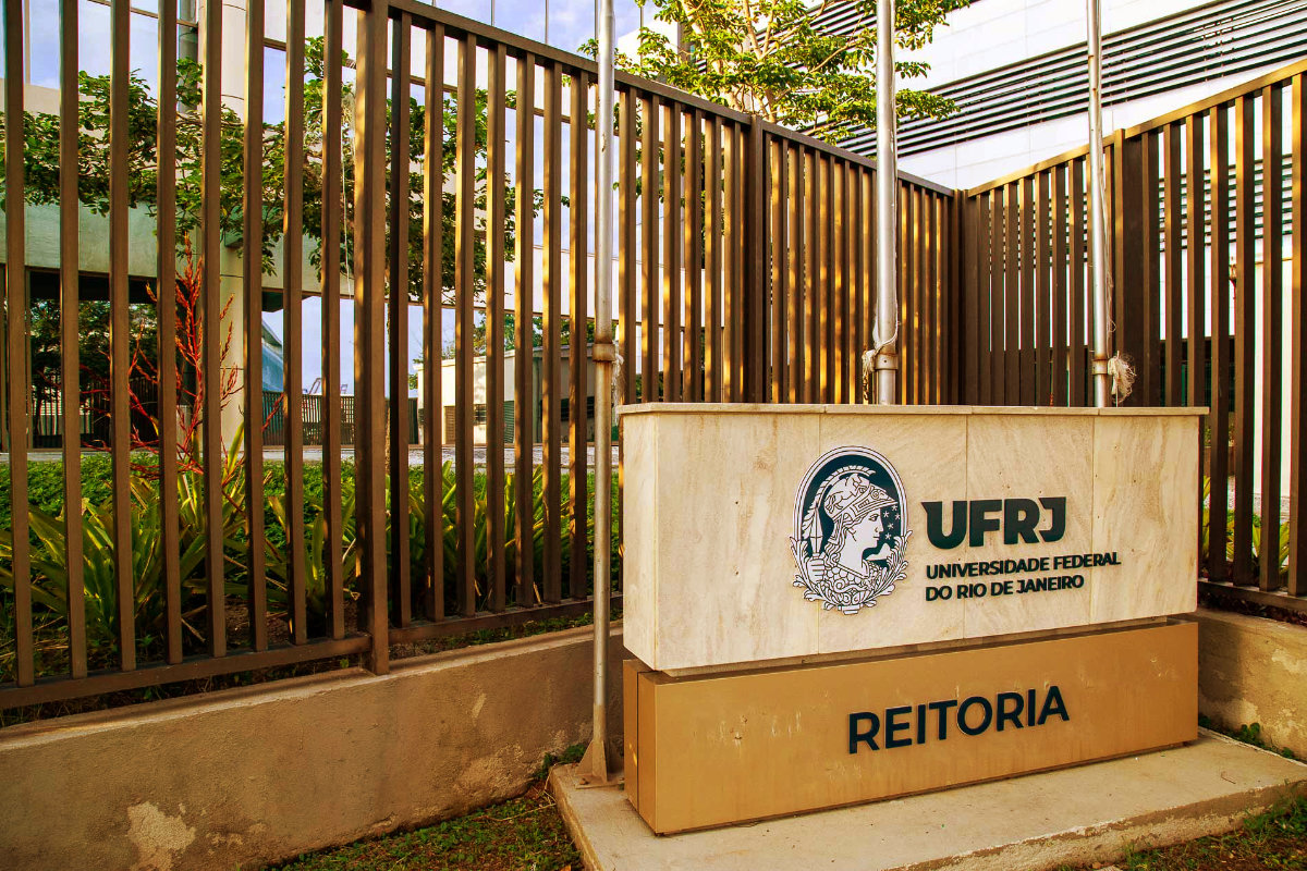 Fotografia mostra, através de grades, parte da fachada do prédio da Reitoria da UFRJ. À frente, há um painel horizontal com a marca completa da Universidade e um letreiro, no qual se lê "Reitoria".